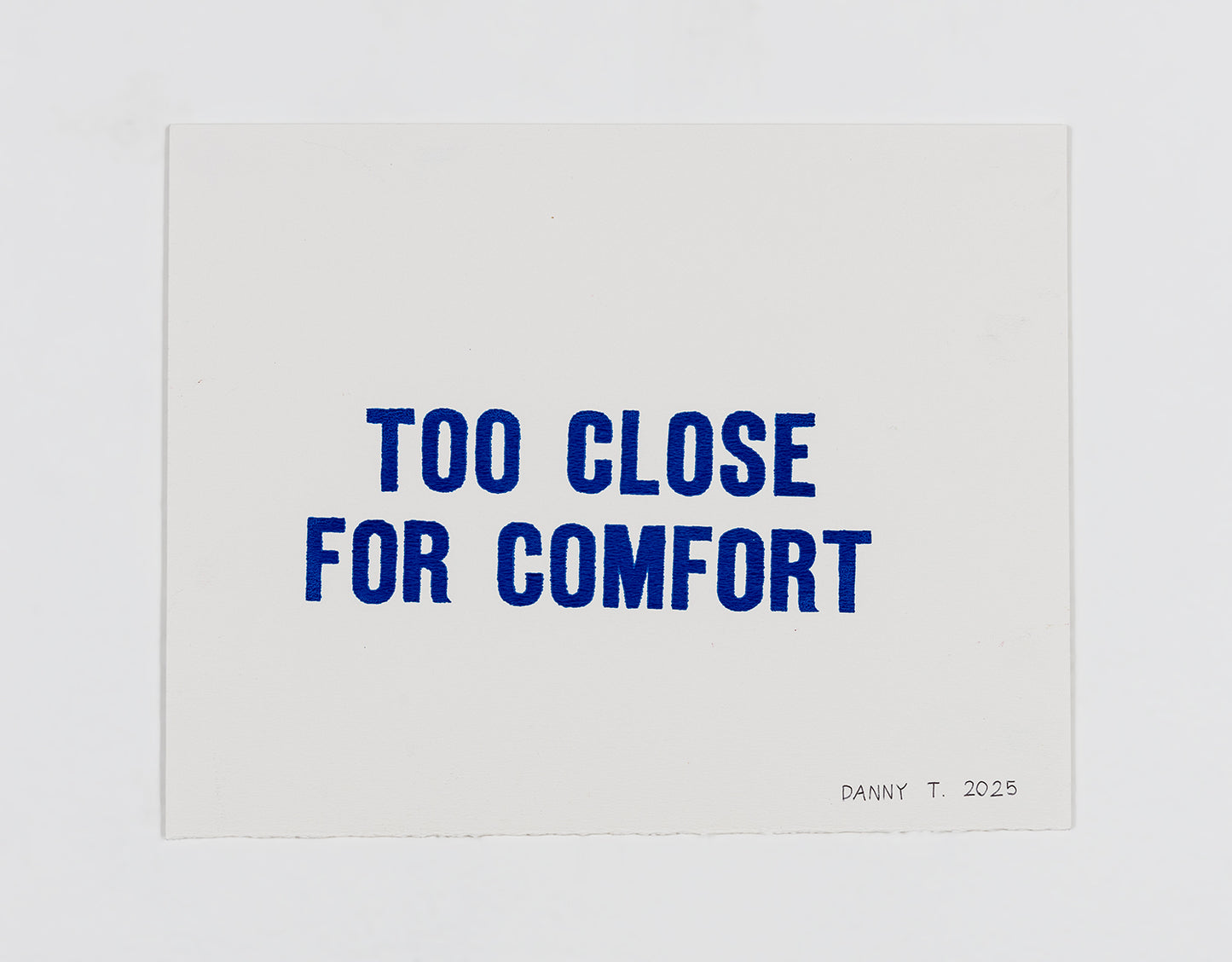 A hand-lettered screen print of two lines of blue text on white paper. The text reads, in all caps: Too close for comfort. 