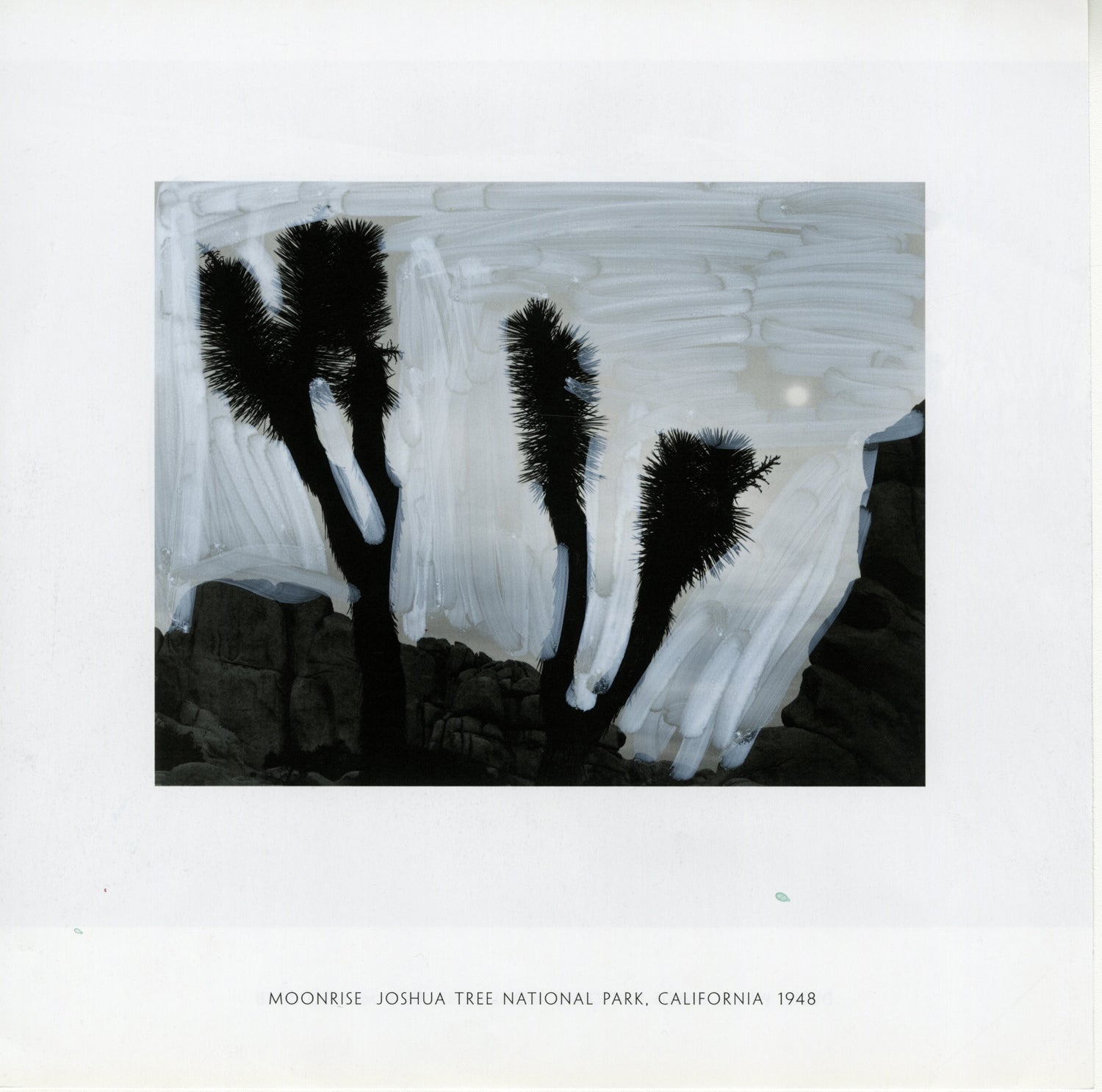 A black and white photograph by Ansel Adams depicting a landscape at Joshua Tree National Park in California, dated 1948. In the center are the silhouetted forms of three Joshua trees with spiky leaves set against the sky. The sky was altered with white acrylic marker, showing a textured, swirling pattern, partially obscuring details and creating an ethereal effect. The moon is faintly visible in the background. The lower portion of the image illustrates rocky terrain, emphasizing the iconic desert scenery.