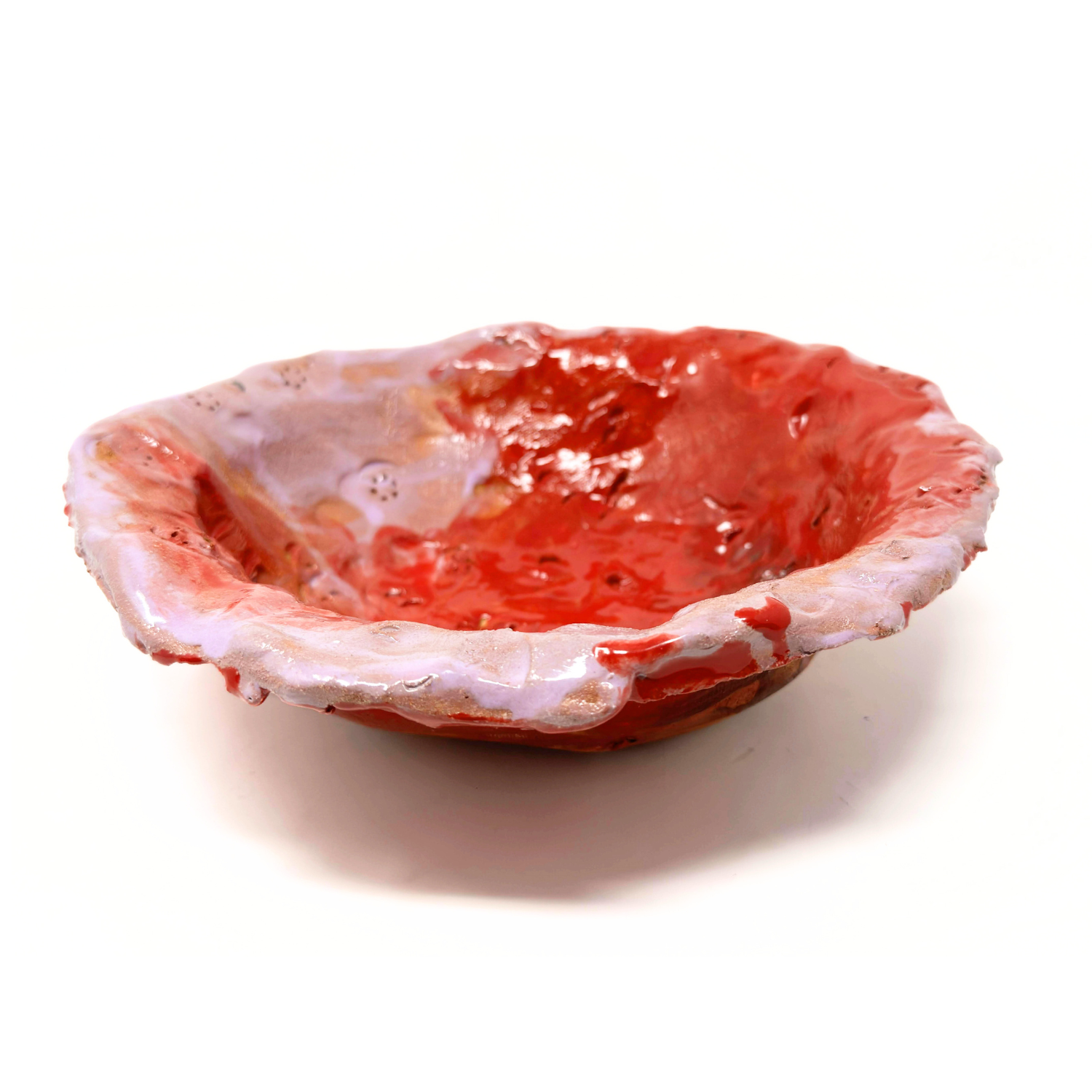 The same hand built ceramic bowl, glazed red and pink, and photographed from the side. The rim of the bowl has areas of sparkly gold layered over the pink and red. The bowl is made by pressing clay into a plaster mold, and the edges maintain the lumpy texture of the clay and the thick glaze. 