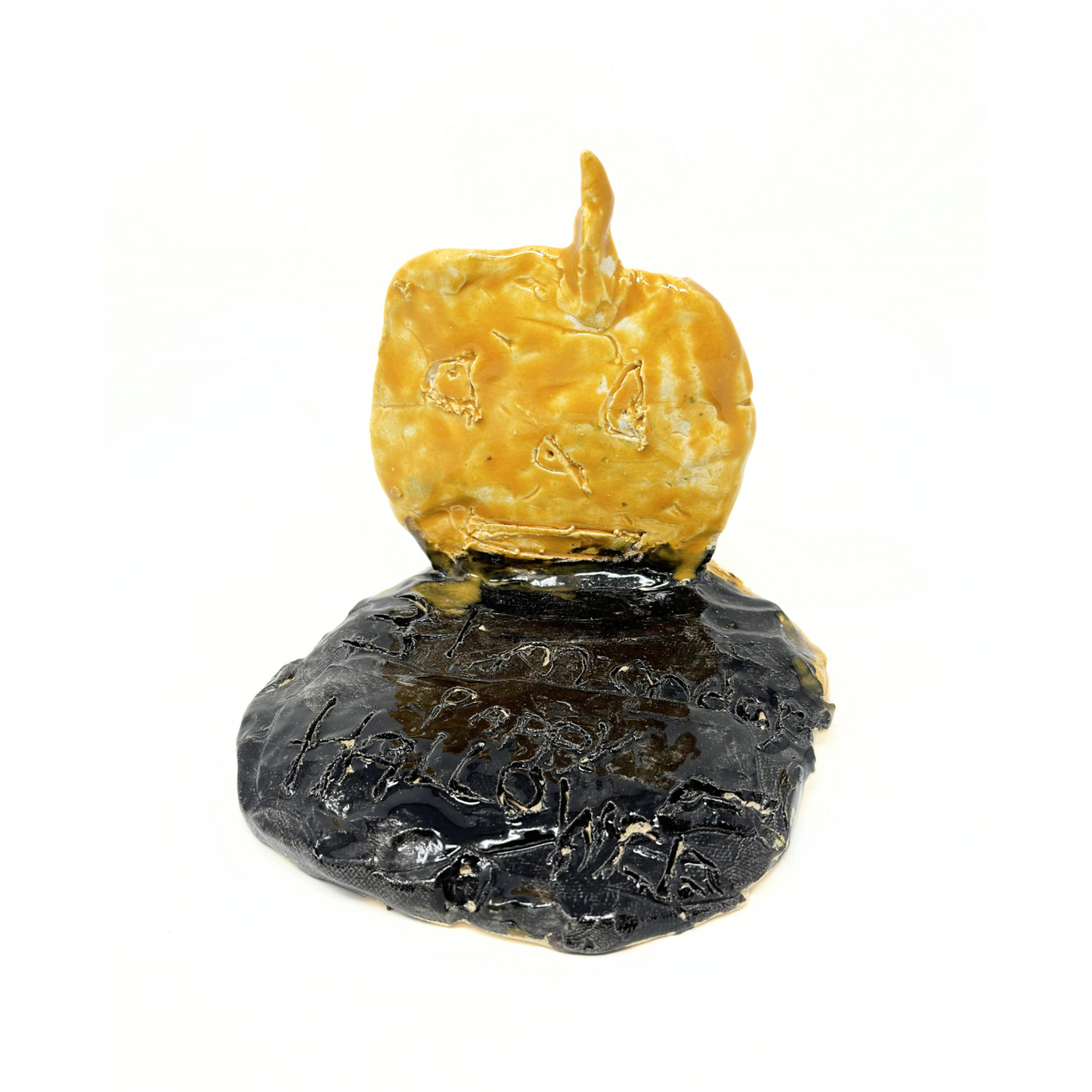 A small hand built ceramic sculpture of an orange jack o lantern pumpkin sitting on a round black base. The jack o lantern has 2 triangle eyes, a triangle nose, and a wide smiling mouth etched on its head. The black base has words etched onto its surface, which read: 
"31 Monday Happy Halloween." All of the glaze used on the sculpture is glossy and shiny. 