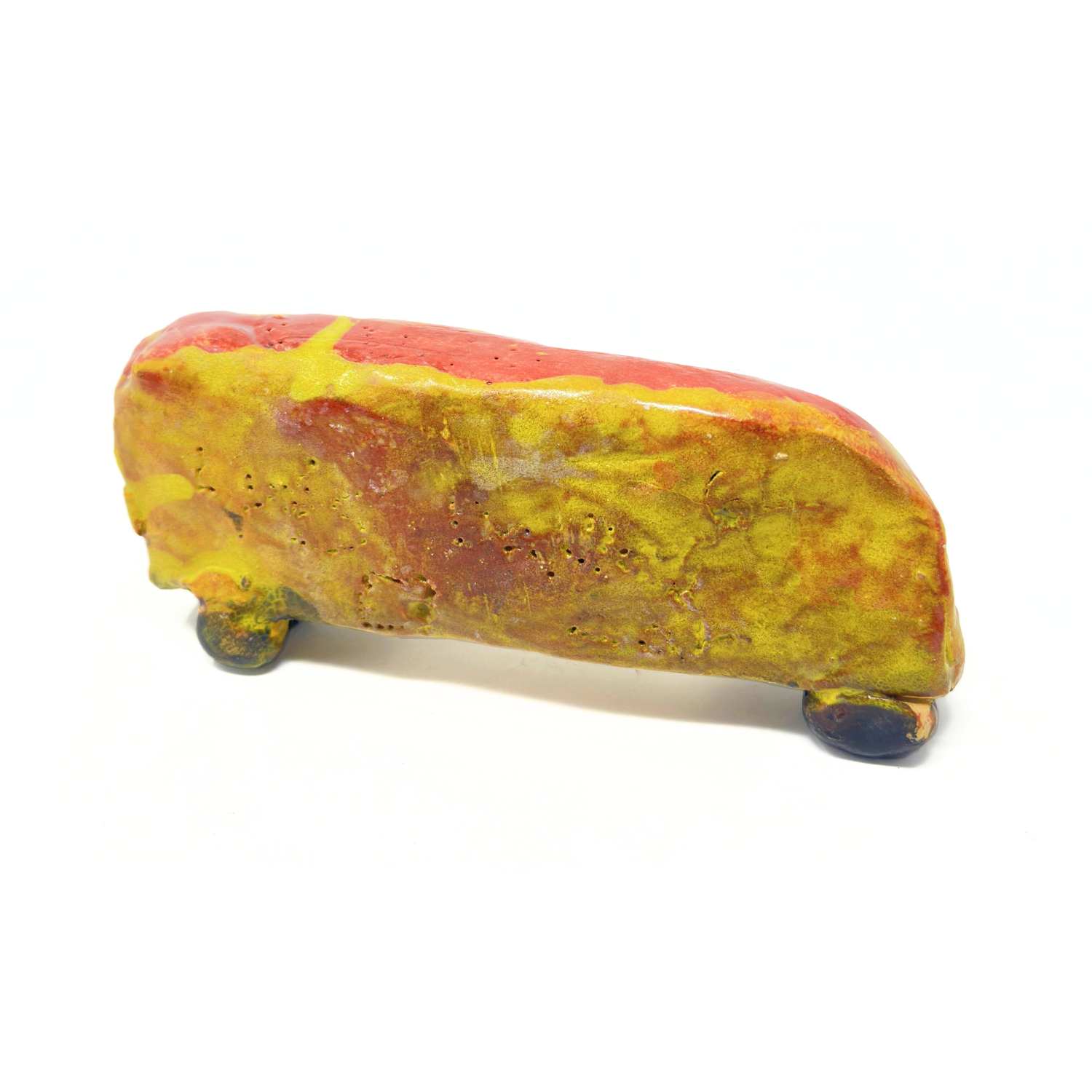 a small ceramic sculpture of a car. The object is a long rectangle with rounded corners, propped upright on 4 small round wheels. The body of the car is glazed red with layers of shiny yellow. The tires are glazed black. The artist has stippled the surface with letters and shapes, some of which spell out his name, "LUIS"