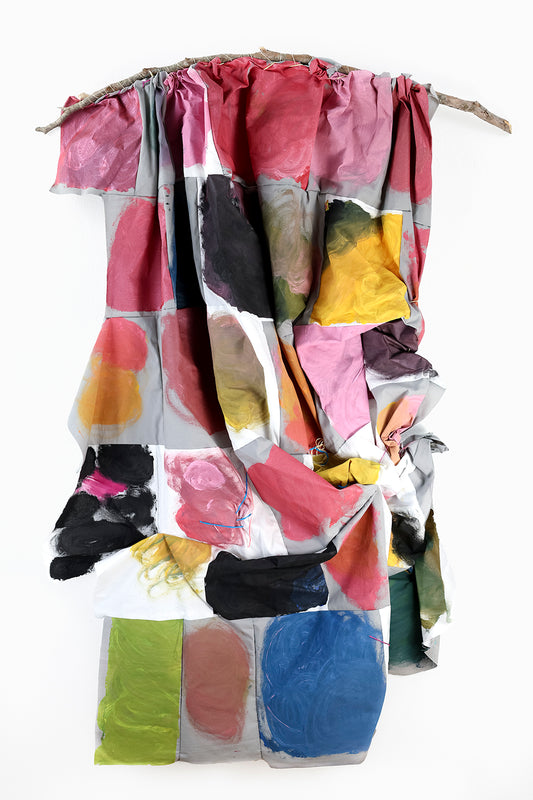 A textile artwork hanging from a tree branch. The fabric is draped unevenly and consists of a patchwork of colorful squares and rectangles. Each section shows a variety of colors, including pink, red, black, yellow, blue, green, and orange, with brushstroke textures. The draping creates folds and shadows, giving the piece a three-dimensional quality. The branch is visible at the top, serving as the anchor for the fabric.