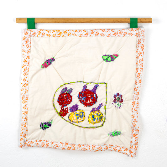 A small square quilt that hangs from a wooden rod with green loops. The fabric is off-white with an orange and yellow leaf-patterned border. Centered on the fabric is an abstract embroidery piece featuring various colored shapes within a green-bordered area. There are red, purple, and yellow circular shapes with textured stitching. Scattered around this central design are small embroidered elements, including colorful leaf-like and floral shapes. 