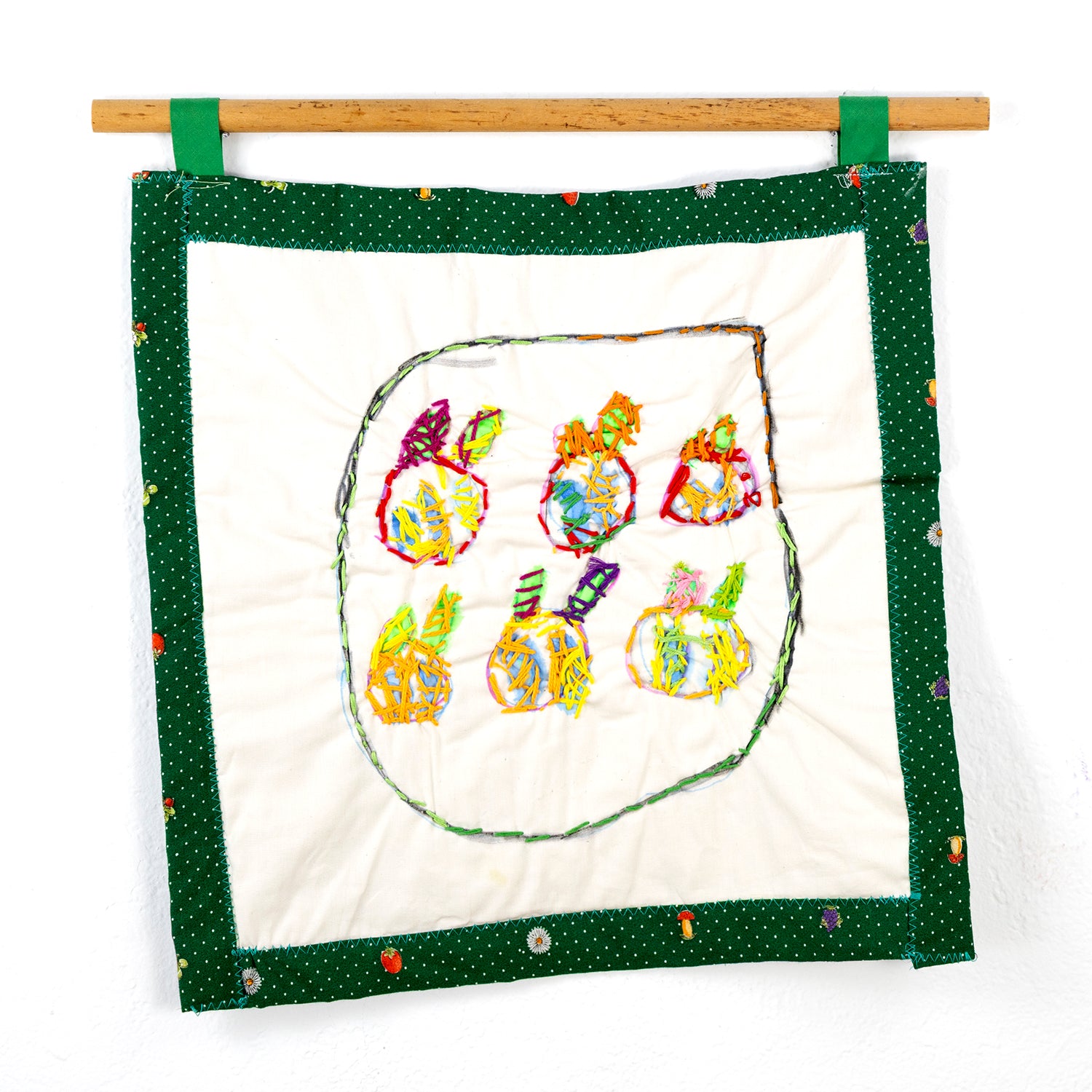 A small square quilt that hangs from a wooden rod with green loops. The quilt features a central design of six stylized fruit-like objects, each crafted with multicolored threads and outlined with vibrant stitching. The edge of the piece has a green border with small white polka dots, adorned with occasional fruit and flower motifs.