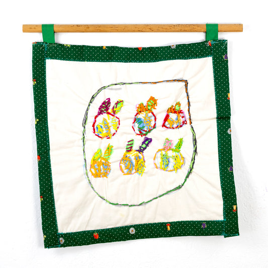 A small square quilt that hangs from a wooden rod with green loops. The quilt features a central design of six stylized fruit-like objects, each crafted with multicolored threads and outlined with vibrant stitching. The edge of the piece has a green border with small white polka dots, adorned with occasional fruit and flower motifs.