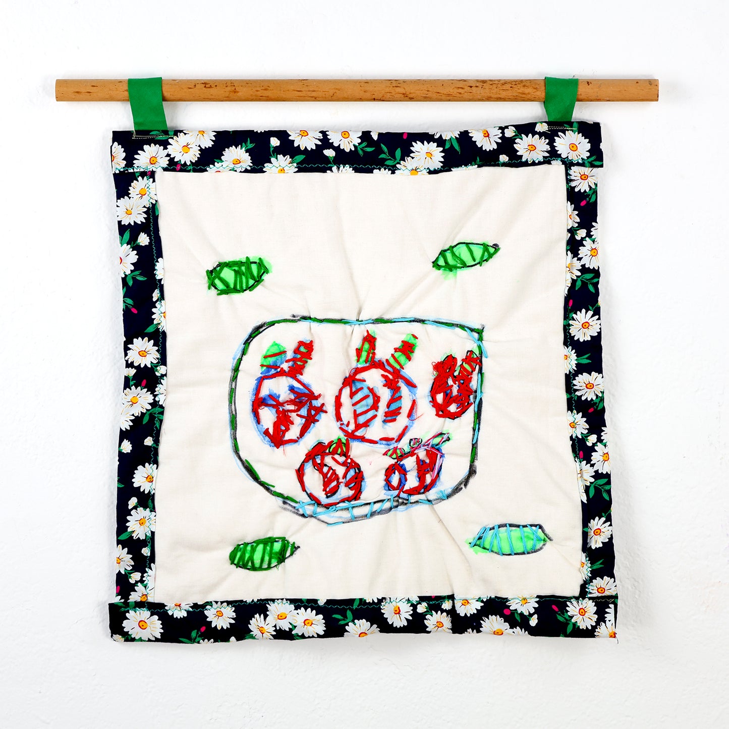 A small square quilt that hangs from a wooden rod with green loops. The centerpiece is a square piece of light beige fabric featuring a stitching of a bowl filled with shapes resembling round fruits, done in vibrant red, blue, and green thread. Surrounding the bowl are green leaf-like shapes embroidered in green thread. The beige fabric is bordered with a black fabric adorned with a white daisy pattern.