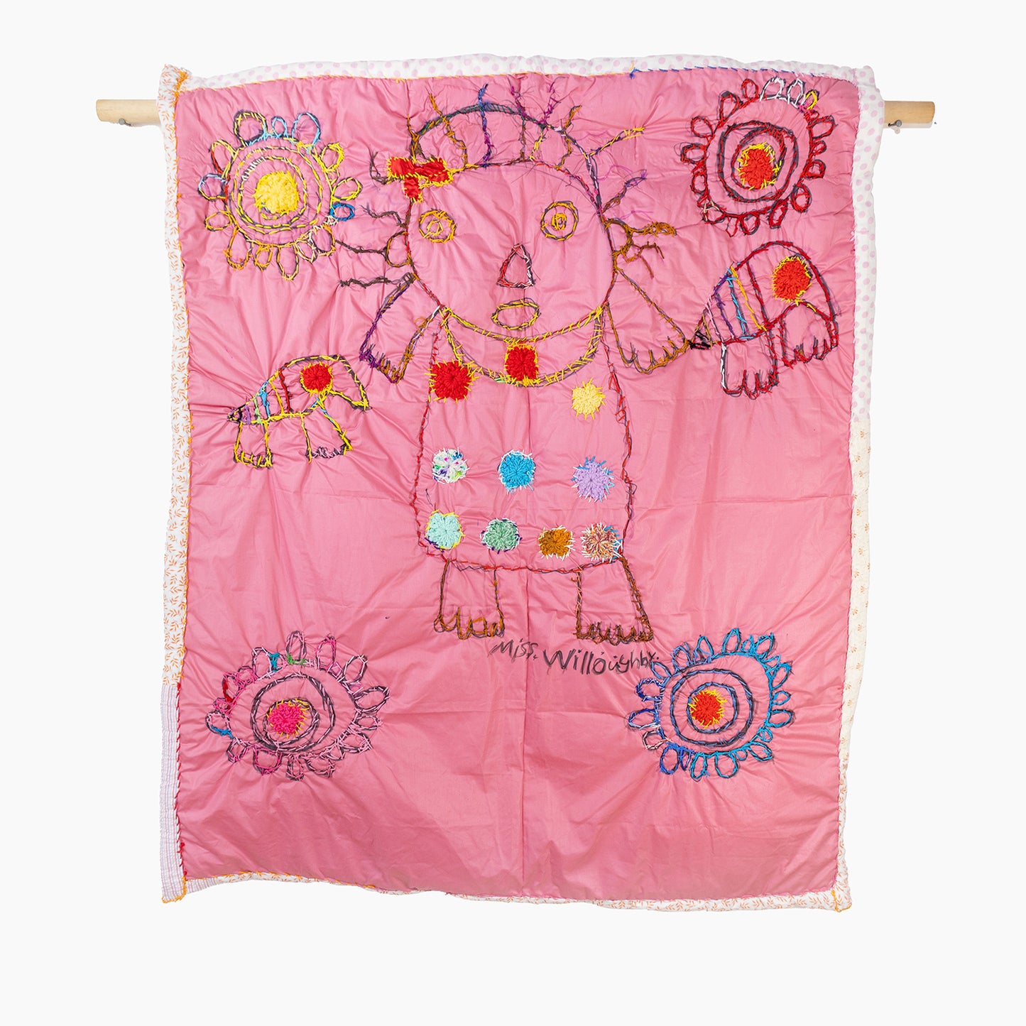 A pink quilt with colorful embroidered designs. In the center, there is an abstract depiction of a person, outlined in brown thread with multicolored hair and eyes. The person is adorned with various circular and floral patterns stitched in vivid reds, greens, blues, yellows, and oranges across their clothing. Surrounding the central figure are several intricate, circular floral motifs, each with a unique combination of colors and styles. The quilt is bordered with a subtle floral or dotted pattern.