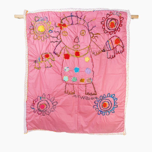 A pink quilt with colorful embroidered designs. In the center, there is an abstract depiction of a person, outlined in brown thread with multicolored hair and eyes. The person is adorned with various circular and floral patterns stitched in vivid reds, greens, blues, yellows, and oranges across their clothing. Surrounding the central figure are several intricate, circular floral motifs, each with a unique combination of colors and styles. The quilt is bordered with a subtle floral or dotted pattern.