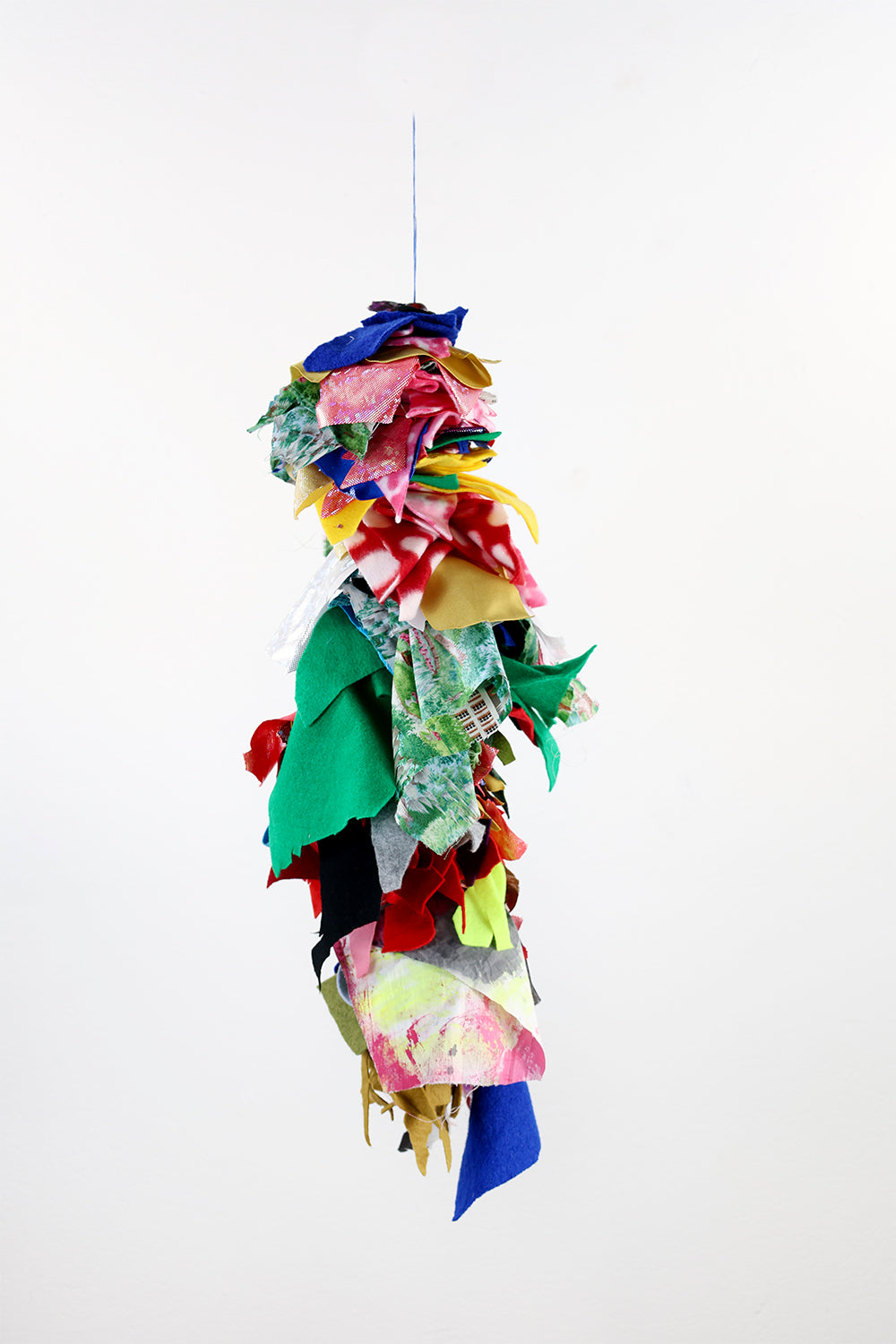 A vibrant, hanging fiber sculpture composed of various fabric scraps. These fabric pieces are layered and knotted onto a vertical string, creating a cascading effect. The fabrics are diverse in color, including blue, red, green, yellow, and multicolored patterns. The composition has an organic, flowing appearance as the fabrics are loosely arranged, with some hanging down freely.