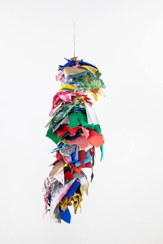 A vibrant, hanging fiber sculpture composed of various fabric scraps. These fabric pieces are layered and knotted onto a vertical string, creating a cascading effect. The fabrics are diverse in color, including blue, red, green, yellow, and multicolored patterns. The composition has an organic, flowing appearance as the fabrics are loosely arranged, with some hanging down freely.