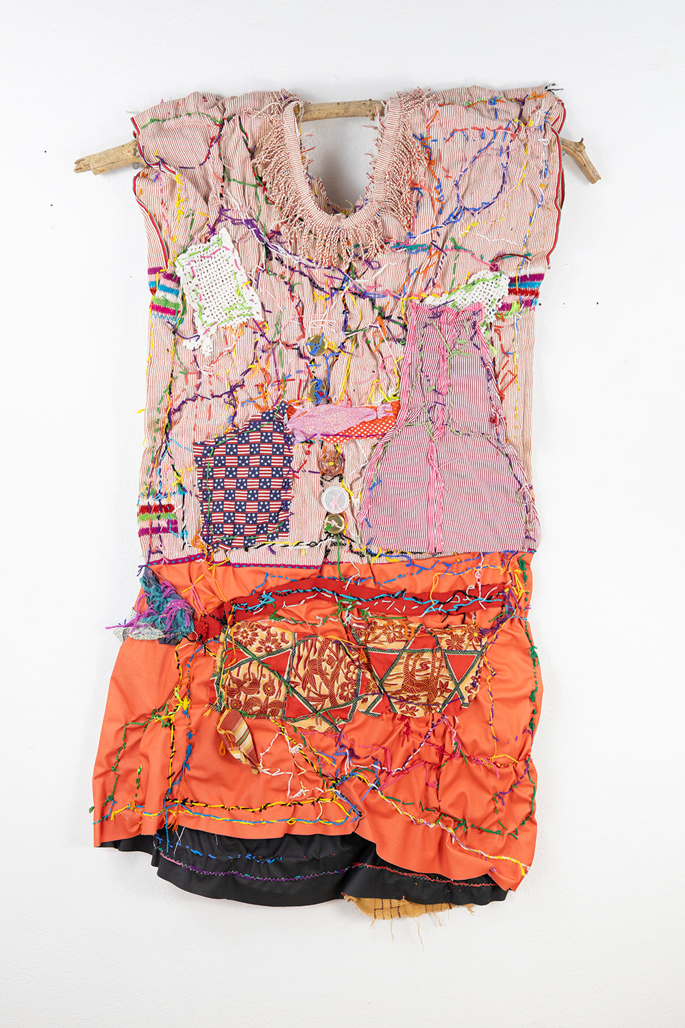 A colorful textile artwork hanging on a wall. The piece is constructed from various fabrics and materials, creating a mixed-media effect. The foundation is a garment-like shape draped from a horizontal stick. The entire piece is adorned with colorful threads that intertwine across the surface, creating an intricate, textured appearance.