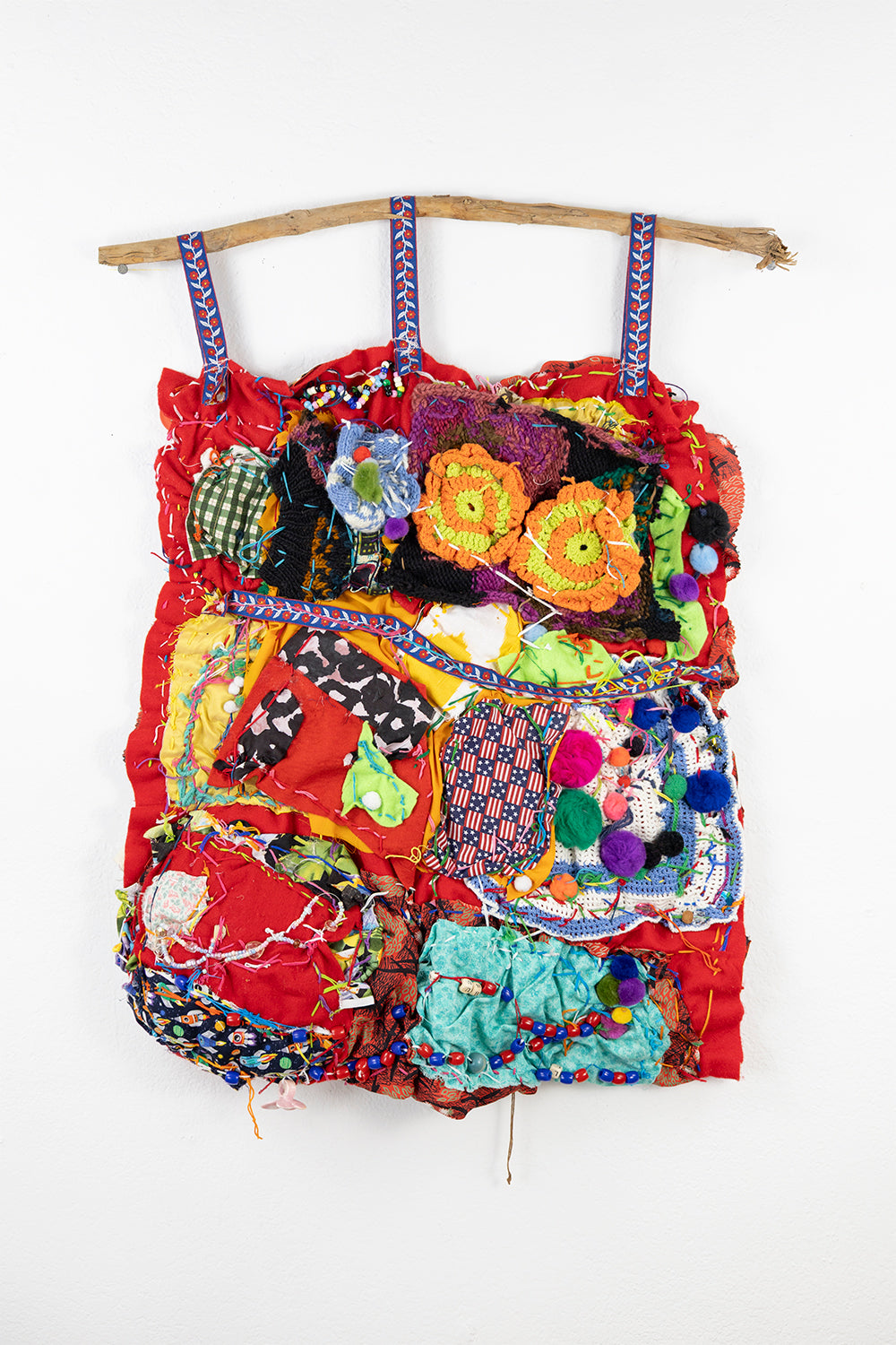 A vibrant textile piece consisting of various colorful fabrics and materials layered and stitched together. It hangs from a curved wooden branch at the top, fastened with three blue fabric loops. The background is a bright red fabric, over which numerous patches of different textures and patterns are sewn. There are decorative elements like ribbon, colorful beads, pom poms, and tangled threads interwoven throughout, creating a rich, textured surface