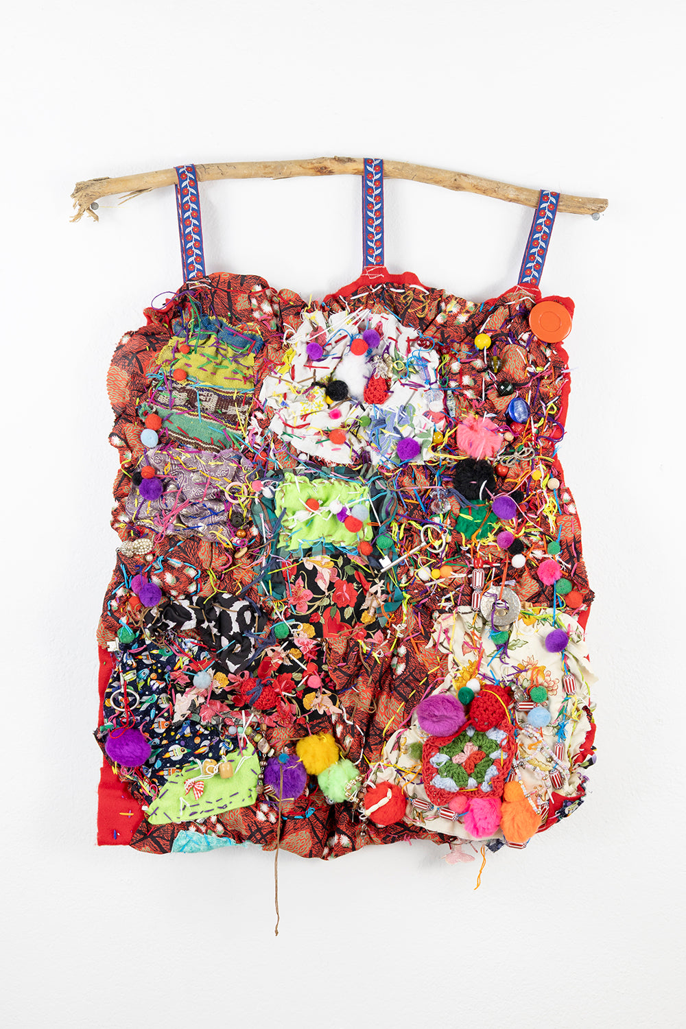 A vibrant textile piece consisting of various colorful fabrics and materials layered and stitched together. It hangs from a curved wooden branch at the top, fastened with three blue fabric loops. The background is a bright red fabric, over which numerous patches of different textures and patterns are sewn. There are decorative elements like ribbon, colorful beads, pom poms, and tangled threads interwoven throughout, creating a rich, textured surface.