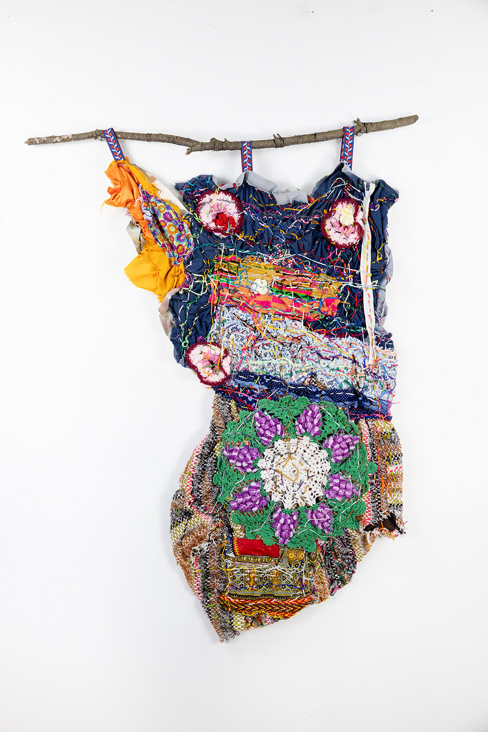 A vibrant textile artwork hanging on a wooden branch affixed to a white wall. The composition includes an array of colorful fabrics and materials. The top section showcases a mixture of dark blue, orange, and patterned fabrics, with red and white circular designs resembling flowers. The lower portion contains a detailed woven or knitted design with a large green and purple floral motif at the center. The entire piece is asymmetrically draped, presenting an organic and irregular shape.
