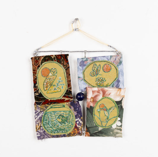 This fabric piece is designed to hang on a wall. The piece has a silver structure that holds up the fabric. The fabric is in four quadrants, connected through a bead sewn into the middle. Each quadrant is a different pattern and contains embroidery of animals and floral elements.