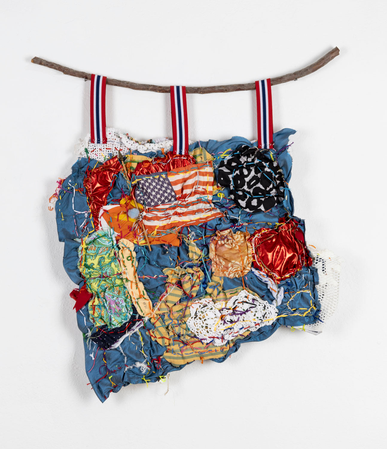 A textile collage on fabric, hanging from a branch, photographed on a white wall. Different swatches of patterned fabric are stitched to a blue base. A mini American flag with a wooden post is stitched near the top. Other swatches feature cheetah, lace, paisley, or striped patterns. A rainbow colored running stitch meanders across the design. 