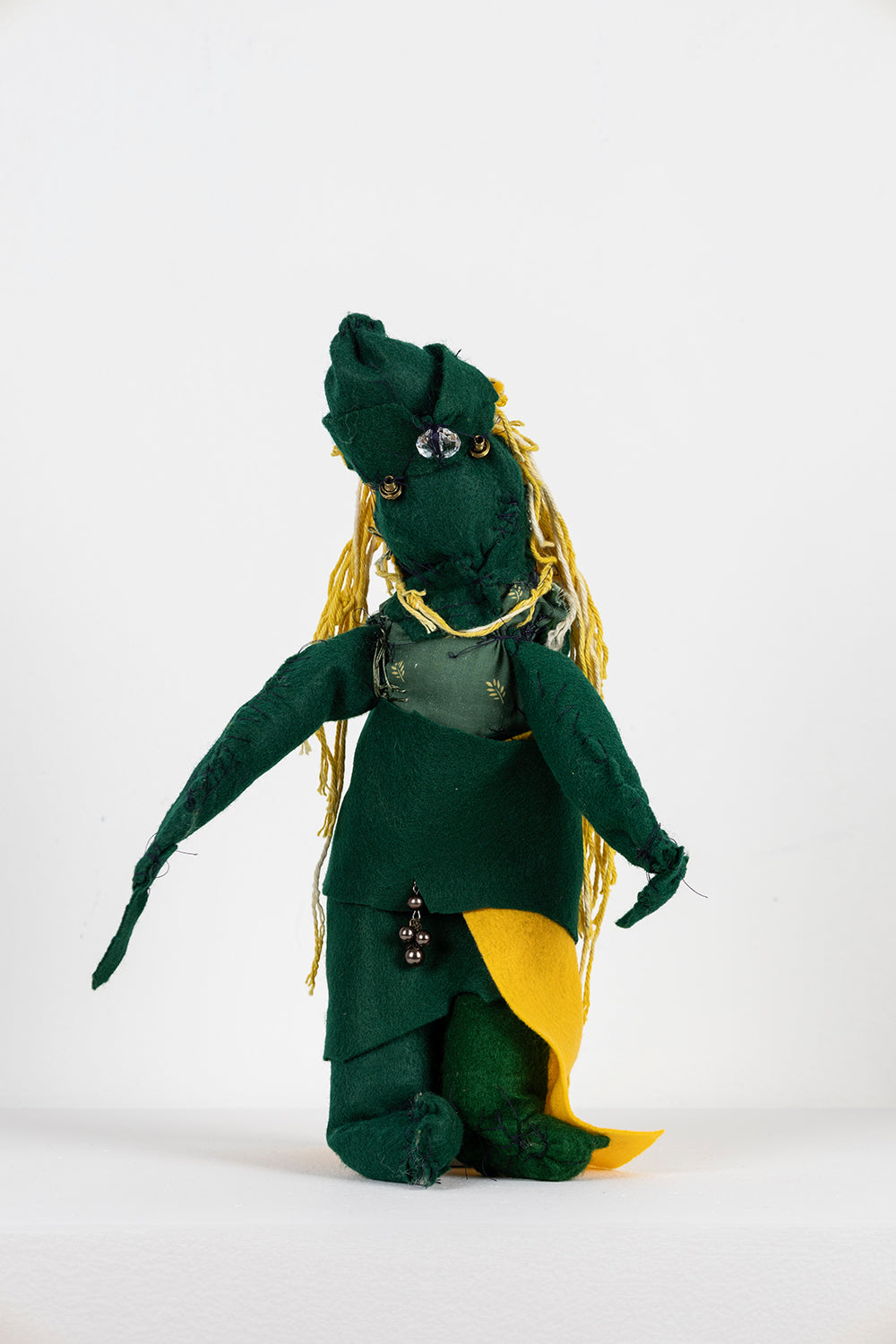 A small soft sculpture made with felt, cotton fabric, yarn, and beads. The sculpture resembles a figure standing upright with both arms gently outstretched. It has yellow yarn cascading down its back like long hair, and beads stitched to its head, positioned like eyes and a nose. From the front, the figure is clad in dark green fabric, but a bit of a yellow felt cape peeks out near their legs. 