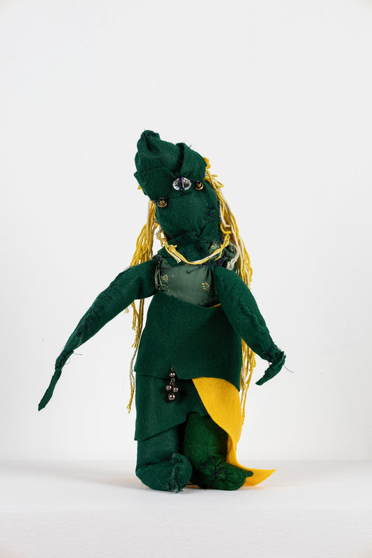 A small soft sculpture made with felt, cotton fabric, yarn, and beads. The sculpture resembles a figure standing upright with both arms gently outstretched. It has yellow yarn cascading down its back like long hair, and beads stitched to its head, positioned like eyes and a nose. From the front, the figure is clad in dark green fabric, but a bit of a yellow felt cape peeks out near their legs. 