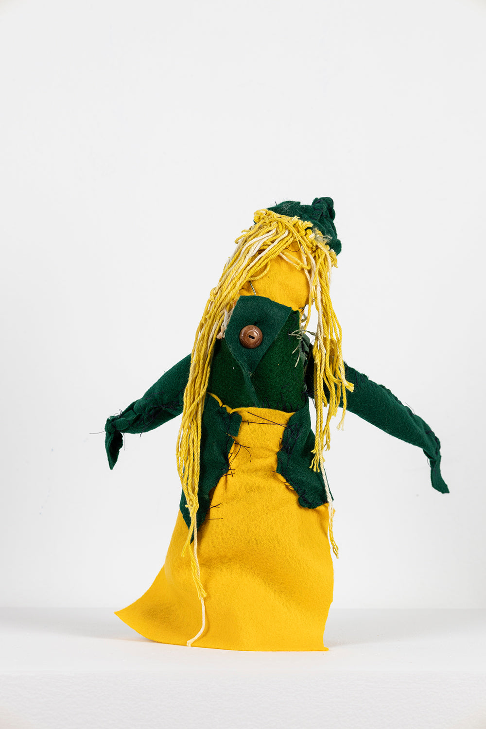 The same sculpture photographed from the back. From this angle, most of the fabrics are bright yellow and dark green. There is a brown wooden button fastening the fabrics together near the character's neck. 