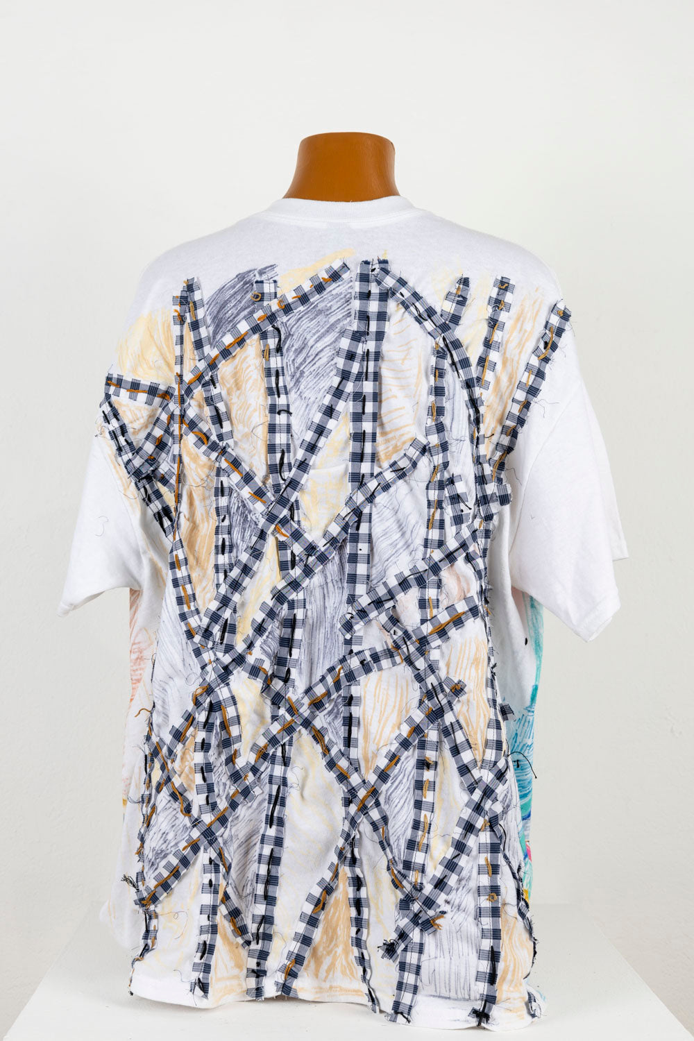 The same white tee shirt, photographed from the back. On this side, the artist has stitched strips of blue and white checkered fabric in a pattern of intersection vertical and diagonal lines. The spaces between each strip have been colored with fabric marker in black, grey, or yellow. 