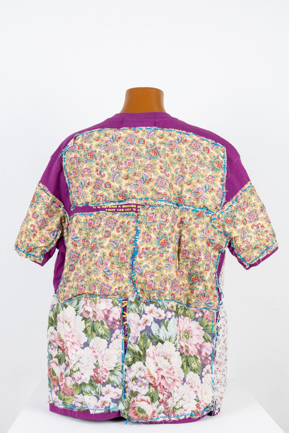 The same purple teeshirt, photographed from the back. Floral swatches of fabric are stitched over most of the shirt, each with a border of bright blue embroidery thread. The patterns printed on the fabric are small or large flowers, some pale pink and realistic, and some more stylized and rainbow-toned. 