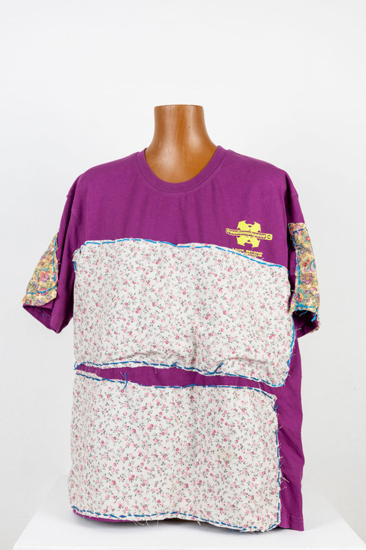 a purple tee shirt with large floral swatches of fabric stitched to the front, back, and sleeves. The fabrics are yellow or white, with printed patterns of tiny pink flowers scattered all over. The teeshirt is photographed on a wooden manikin torso, positioned before a white wall. 