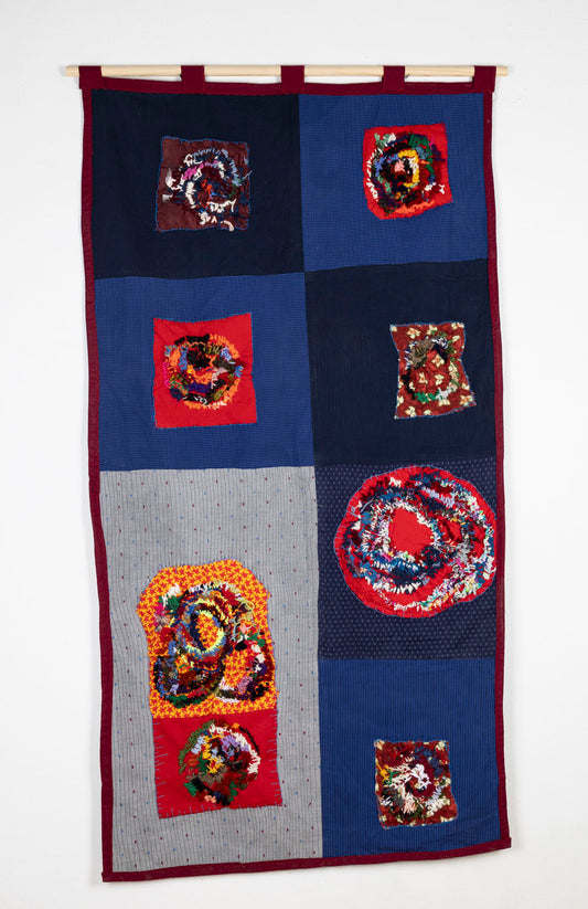 A patchwork embroidered quilt hanging from a wooden dowel photographed against a white wall. Large blue and navy squares of fabric form a 2 by 4 checkerboard. In the center of each square, the artist has appliquéd a red swatch of fabric. Each red swatch has been densely embroidered over with rings of rainbow colored thread, some tracing the imprint of quilting hoops. A thin red band runs around the border of the quilt.