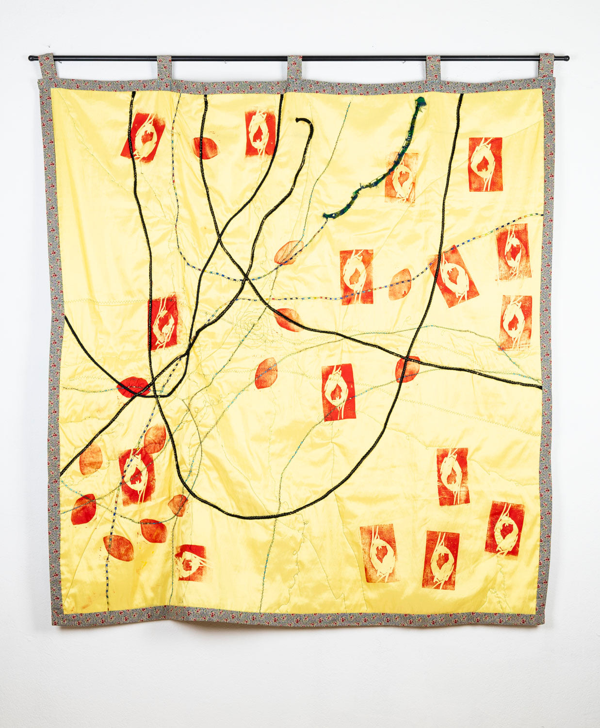 A mixed media textile work: a large square of yellow satin fabric with a thin floral border hangs from a black dowel. Several long synthetic hair braids loop and intersect across the surface, stitched down with green thread. Using red ink, the artist has printed the yellow fabric with a sprawling pattern of different lino-block stamps. Many of these stamps are shaped like petals or eyes. 