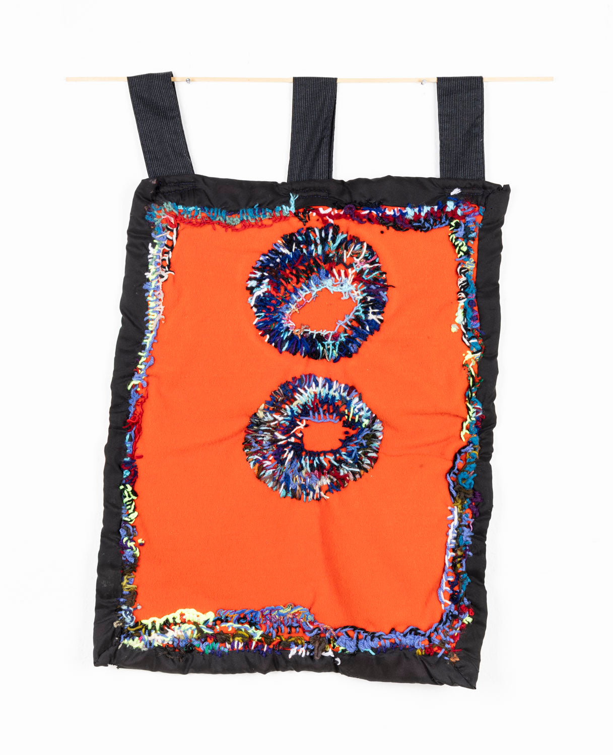 This fiber piece is designed to hang, as shown in the photograph. The base fabric is black, with an orange center. There are two circles embroidered in the middle of the piece, stacked vertically. The circles are a combination of vibrant threads; red, blue, black, green and purple. The same thread colors are also used to connect the black and orange fabrics.