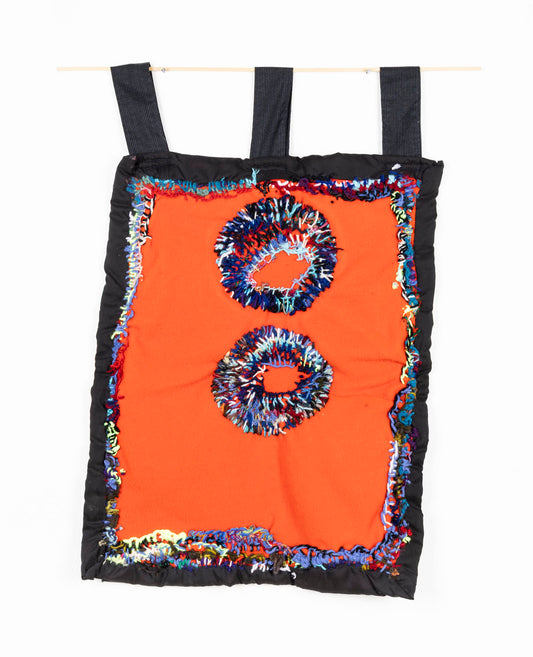 This fiber piece is designed to hang, as shown in the photograph. The base fabric is black, with an orange center. There are two circles embroidered in the middle of the piece, stacked vertically. The circles are a combination of vibrant threads; red, blue, black, green and purple. The same thread colors are also used to connect the black and orange fabrics.