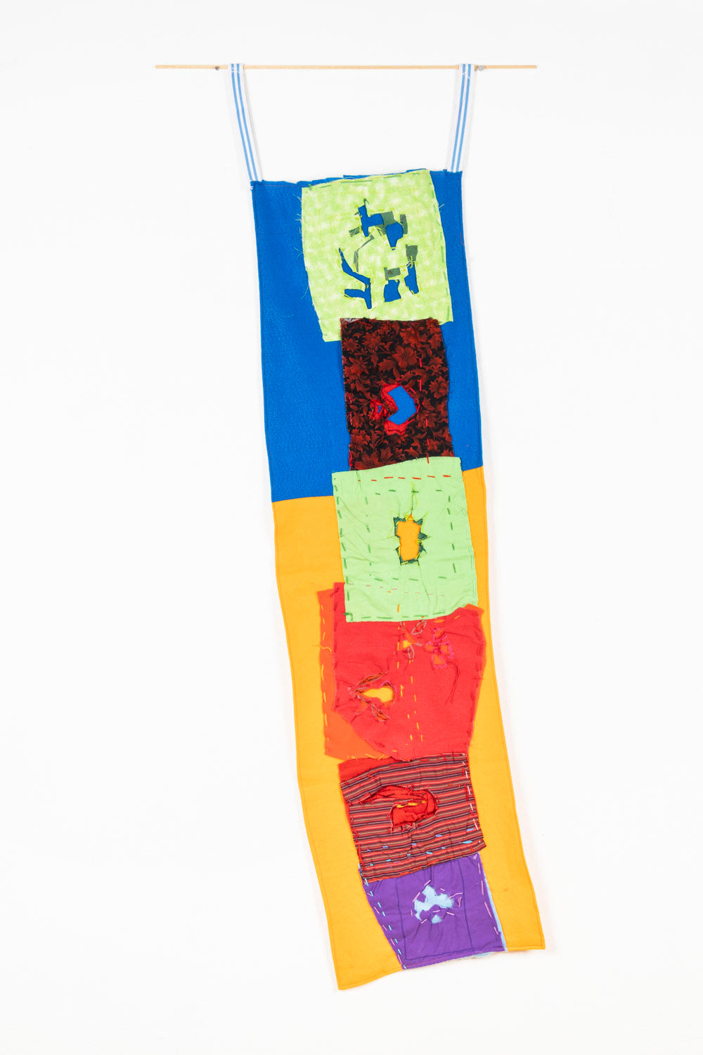 This fabric piece is designed to hang, as shown in the image. There is a collage-like quality to the piece; the background is blue on the top and yellow on the bottom, with patches with varying patterns embroidered on top. The embroidered patches are green, red and purple, that contain cutouts so the background colors are able to shine through.