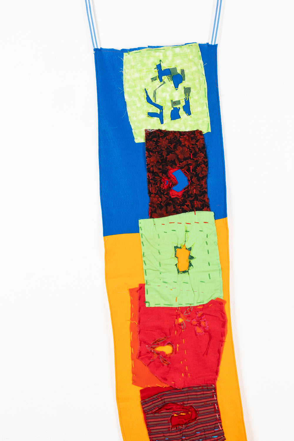 This fabric piece is designed to hang, as shown in the image. There is a collage-like quality to the piece; the background is blue on the top and yellow on the bottom, with patches with varying patterns embroidered on top. The embroidered patches are green, red and purple, that contain cutouts so the background colors are able to shine through.