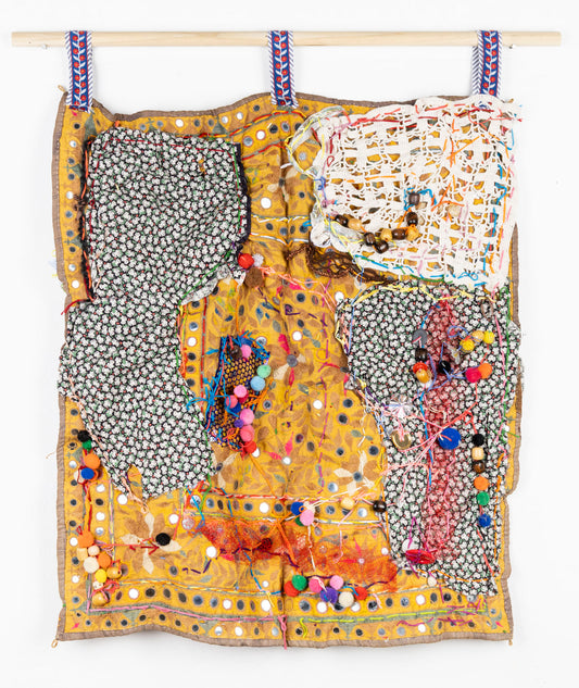 This fabric piece is designed to hang against a wall or in space. The piece has a collage-like quality, with various fabrics and materials overlapping and layered. There is a black and white floral fabric used in the top left and bottom right sections of the piece, with multi-colored yarn and pompoms sewn on top. In the top right corner there is a white, lace fabric that allows you to see yellow fabric underneath. 