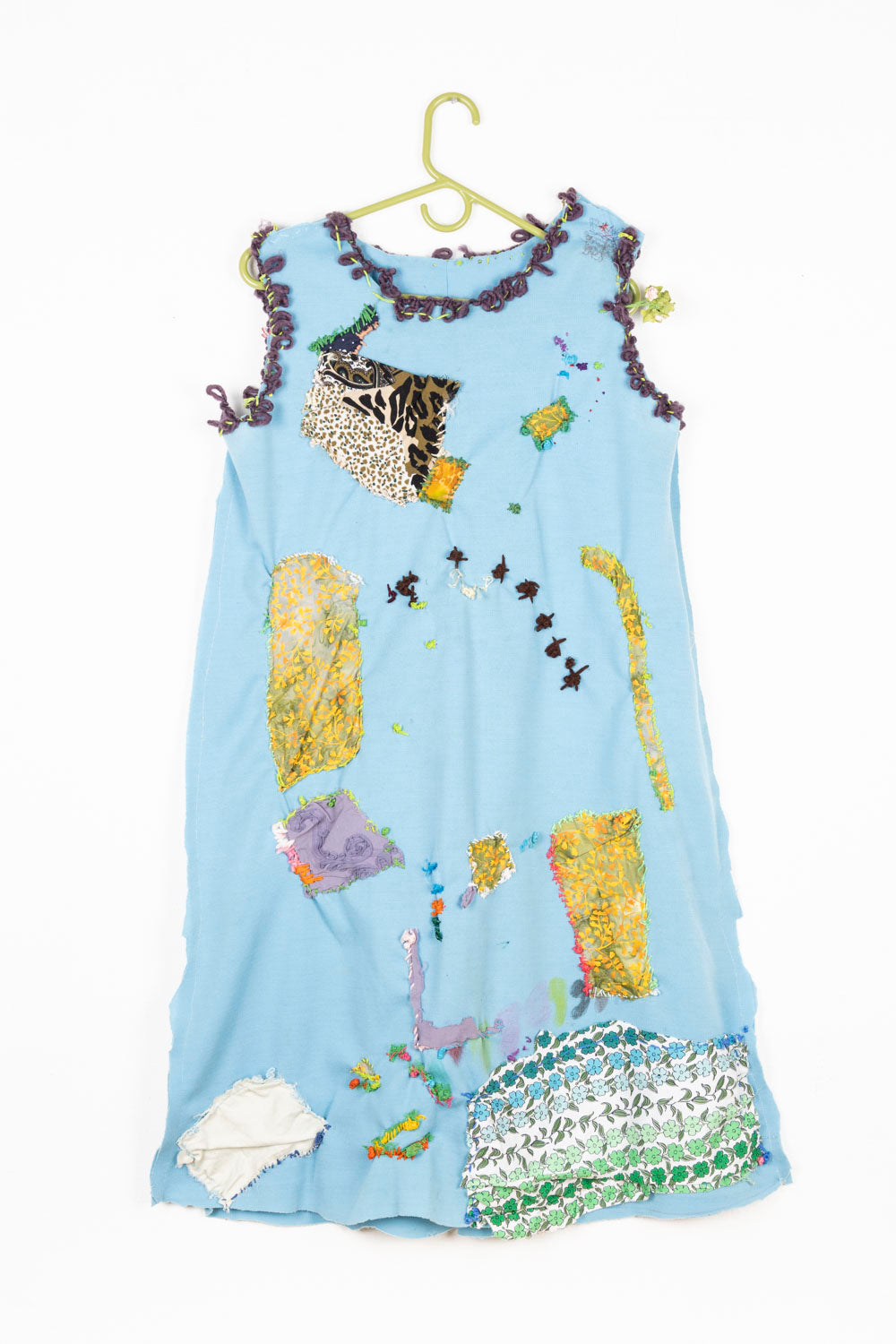 This is a light blue dress with purple embroidery on the collar and the arm holes. The dress also has patches of patterned fabrics embroidered onto the dress. 