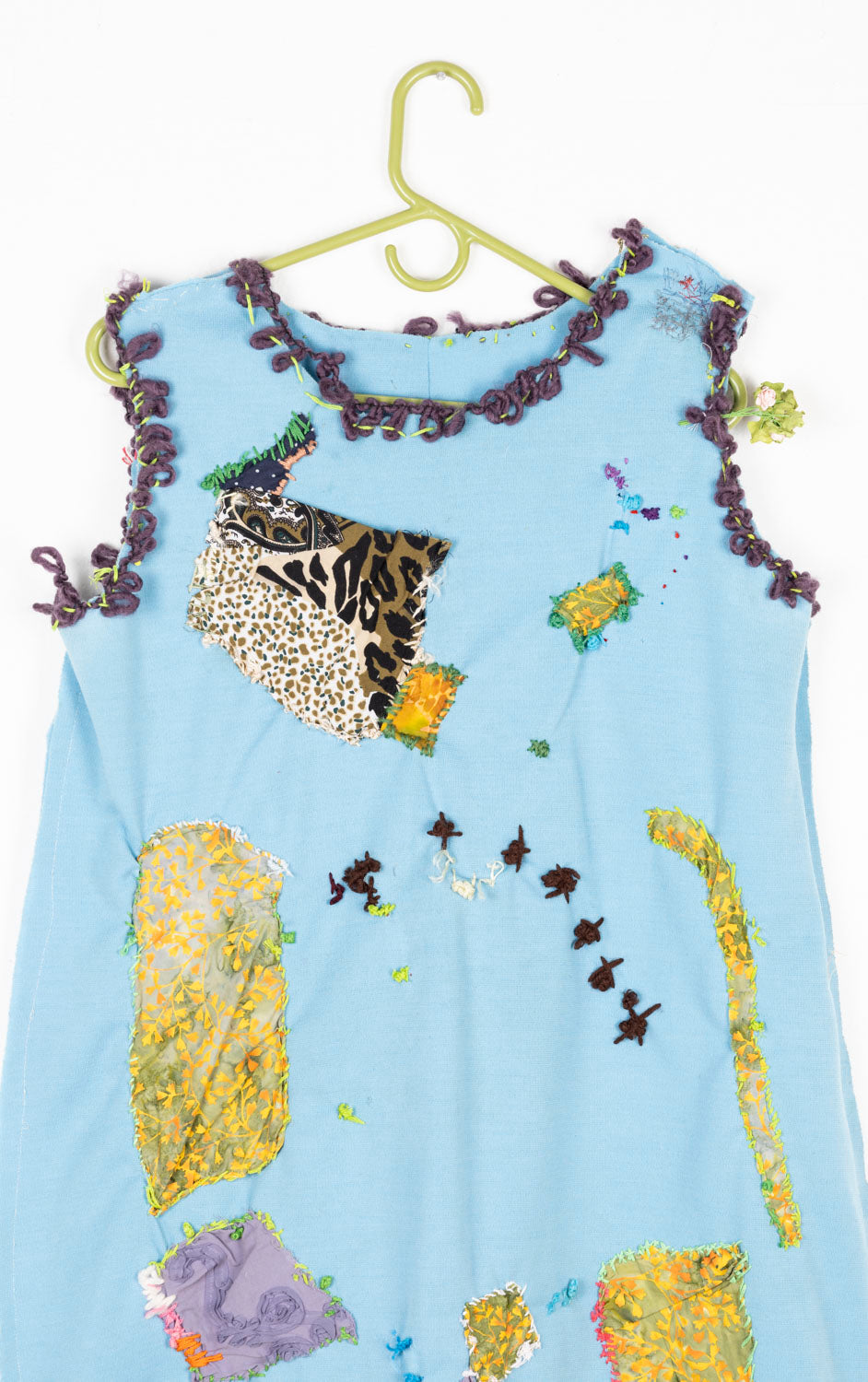 This is a light blue dress with purple embroidery on the collar and the arm holes. The dress also has patches of patterned fabrics embroidered onto the dress. 