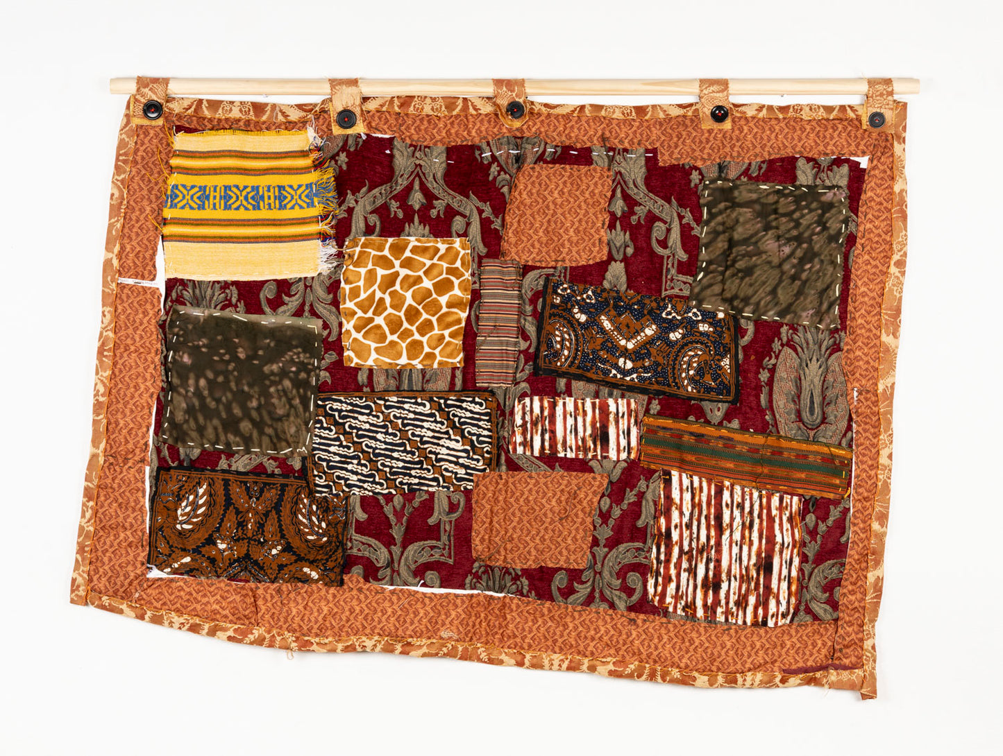 This is a fiber piece hanging on a wooden dowel. The base fabric is orange with a brown floral pattern. On top there are patches with different patterns that are layered and embroidered on with white thread.