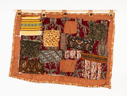 This is a fiber piece hanging on a wooden dowel. The base fabric is orange with a brown floral pattern. On top there are patches with different patterns that are layered and embroidered on with white thread.