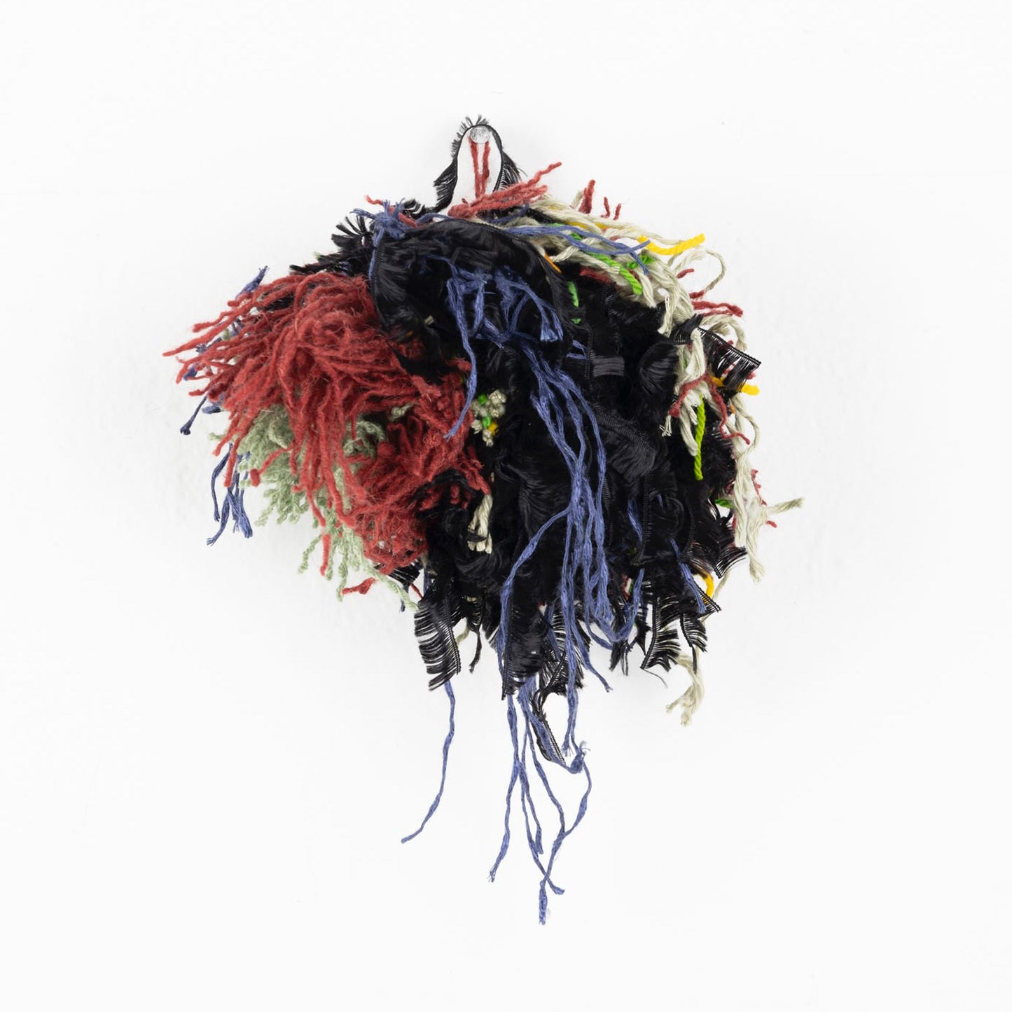 a photo of a colorful yarn pom pom ball: short bits of black, red, green, tan, and blue yarn are knotted together in a round clump. Some of the yarn hangs down in longer feathery strands. 