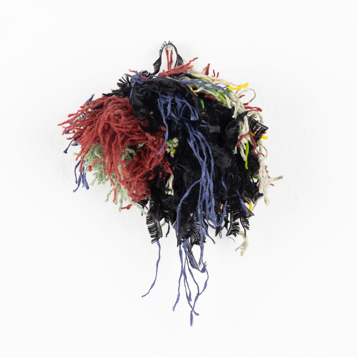a photo of a colorful yarn pom pom ball: short bits of black, red, green, tan, and blue yarn are knotted together in a round clump. Some of the yarn hangs down in longer feathery strands. 