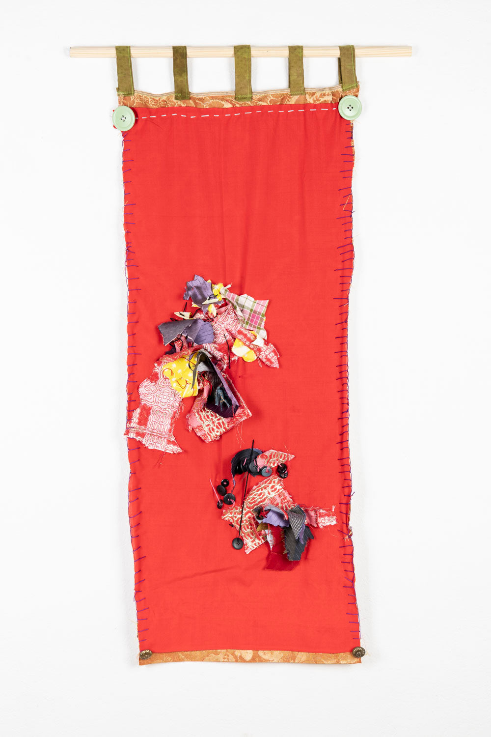 This is a fiber piece hanging on a wooden dowel. The fabric is a bright red, with orange details on the top and the bottom. There are also fabric scraps of various patterns sewn onto the piece. 