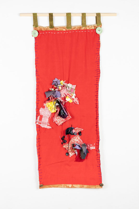This is a fiber piece hanging on a wooden dowel. The fabric is a bright red, with orange details on the top and the bottom. There are also fabric scraps of various patterns sewn onto the piece. 