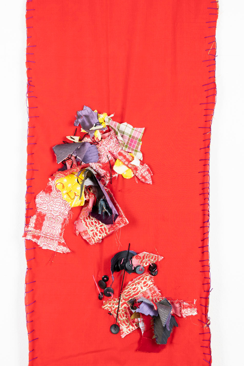 This is a fiber piece hanging on a wooden dowel. The fabric is a bright red, with orange details on the top and the bottom. There are also fabric scraps of various patterns sewn onto the piece. 