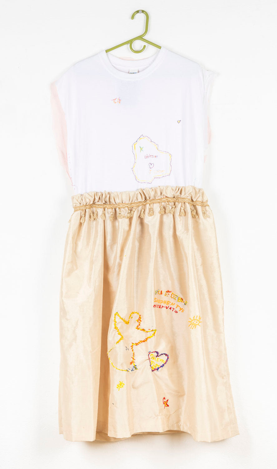 This wearable garment has a white top sewn onto a gold skirt. The waist of the garment is cinched with gold tassels. Both the top and the skirt have embroidered elements; the words 'Liliesa', 'Stephen Tys' and 'Obserato', hearts and the sun. 