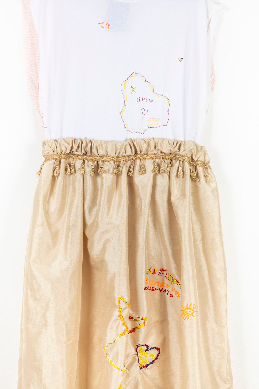 This wearable garment has a white top sewn onto a gold skirt. The waist of the garment is cinched with gold tassels. Both the top and the skirt have embroidered elements; the words 'Liliesa', 'Stephen Tys' and 'Obserato', hearts and the sun. 