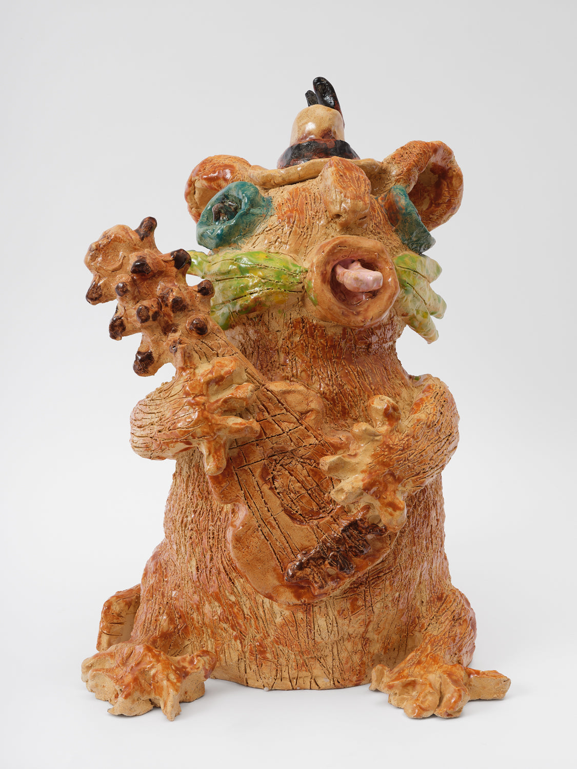 A ceramic cat sculpture, the cat sits up on its hind legs and holds a guitar in its hands. The cat is light brown and has many lines throughout the surface of its body, delineating fur. The cat wears a small hat, has big circular teal eyes, fat green whiskers, and a light pink tongue sticks out of its mouth.