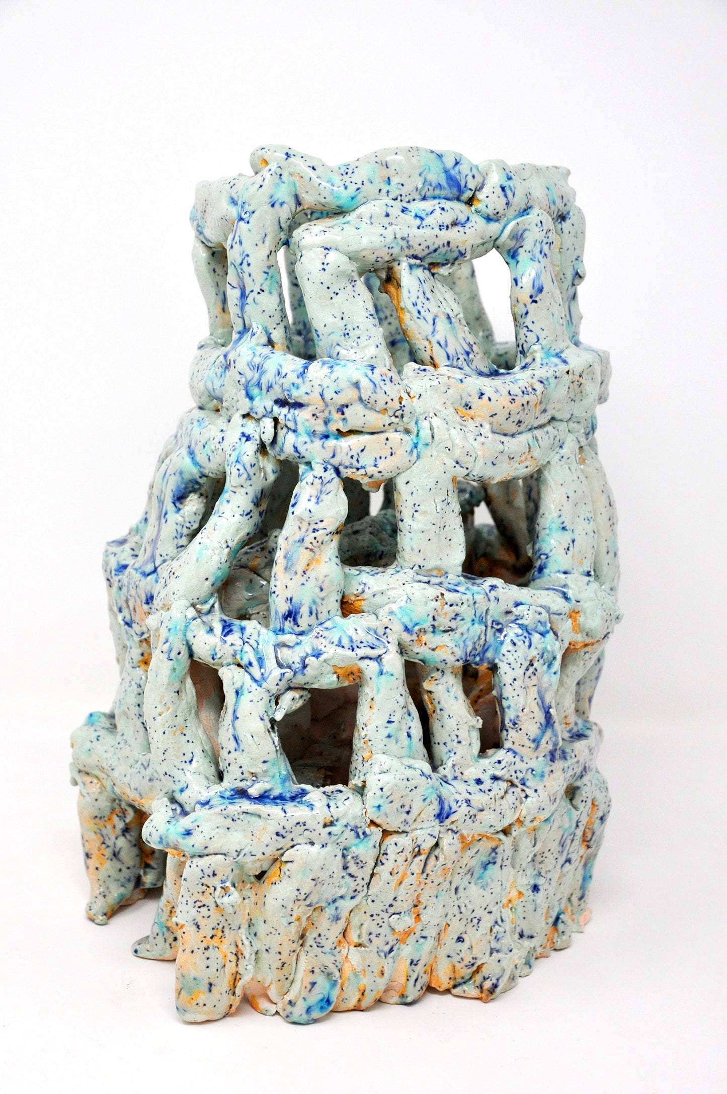 A ceramic sculpture of a tower. It's coated with glossy teal glaze speckled all over with bright royal blue spots. The tower is four tiers tall, wider at the base. It's made of little cylinders: some are stacked vertically and others are wrapped around the tower in horizontal bands. There are gaps between each cylinder through which the white background or dark interior are visible.