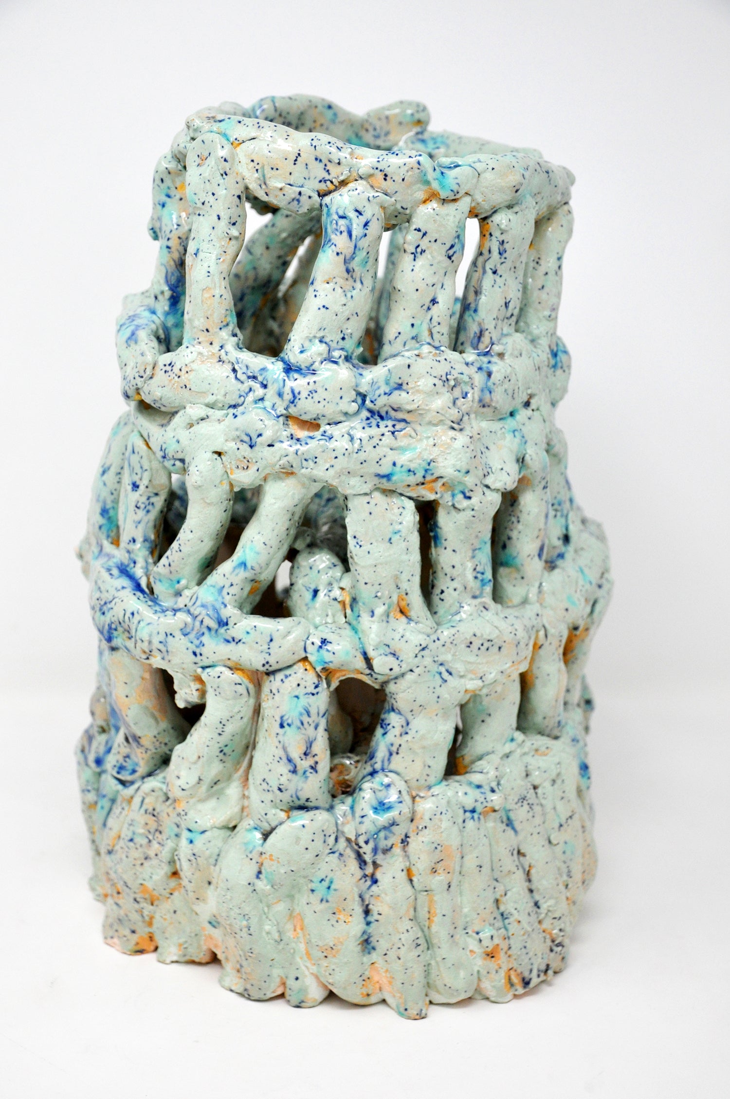 A ceramic sculpture of a tower. It's coated with glossy teal glaze speckled all over with bright royal blue spots. The tower is four tiers tall, wider at the base. It's made of little cylinders: some are stacked vertically and others are wrapped around the tower in horizontal bands. There are gaps between each cylinder through which the white background or dark interior are visible.