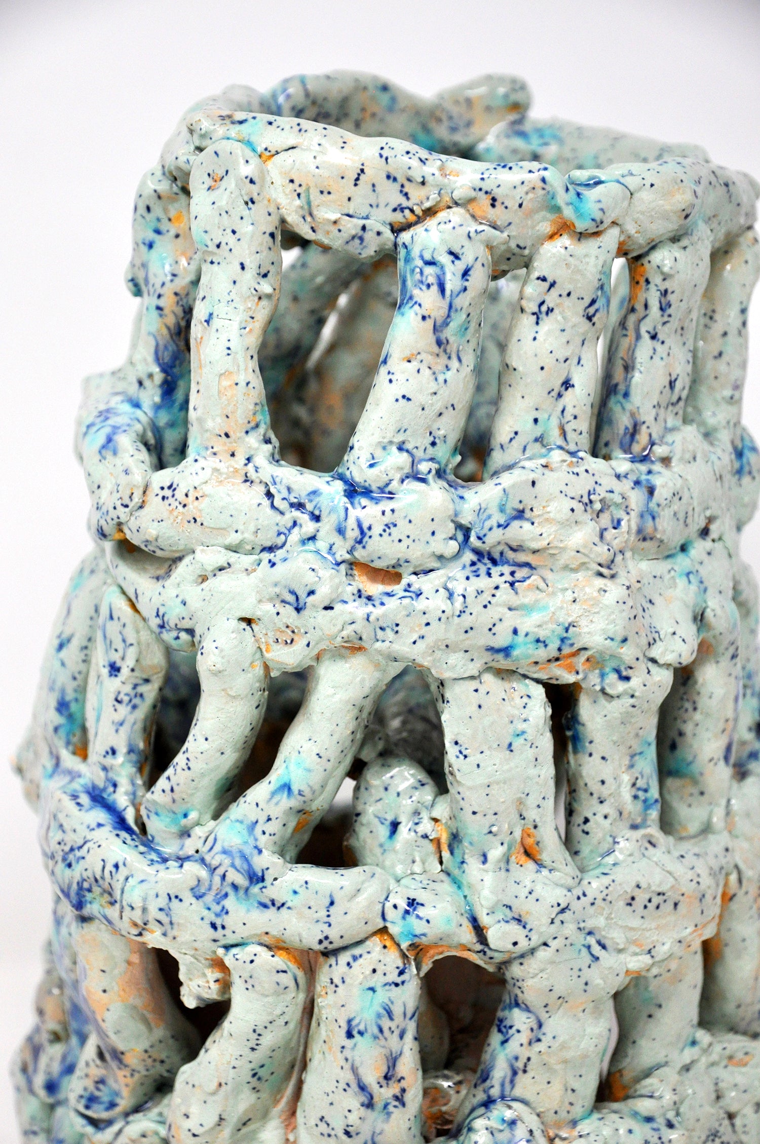 A detail of the top of a ceramic tower sculpture. It's coated with glossy teal glaze speckled all over with bright royal blue spots. The tower is like a woven basket or cage -- it's made of little cylinders: some are stacked vertically and others are wrapped around the tower in horizontal bands. There are gaps between each cylinder through which the white background or dark interior are visible.