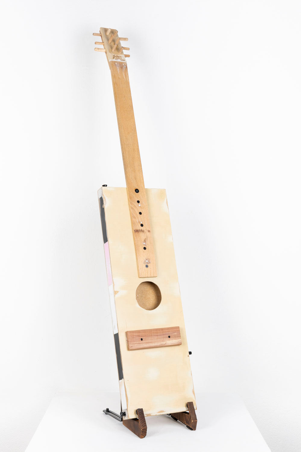 A handmade instrument photographed on a stand on a white pedestal. The instrument is a guitar made from reclaimed wood and metal. The body of the guitar is long and rectangular, and the tuning headstock is lined with little wooden pegs. The artist writes that this guitar sculpture was made as a tribute to his father who encouraged imaginative and creative use of recycled material.  