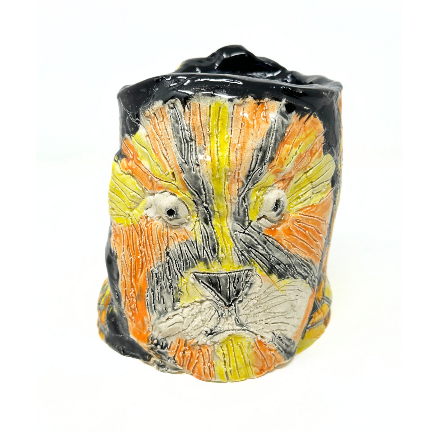 The same ceramic vessel photographed from another angle, opposite of the queen portrait. This side of the vessel has a portrait of a lion face, etched with textured fur lines, and glazed orange, yellow, and black. The lion has two round white eyes with black pupils, a triangular black nose, and a small down turned mouth. The space around the lion head is glazed shiny black. 